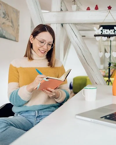 Eine Frau sitzt entspannt an einem Schreibtisch in einem hellen, weihnachtlich geschmückten Zimmer. Sie hält ein Notizbuch in der Hand und schreibt lächelnd hinein. Vor ihr stehen ein Laptop und eine Tasse.