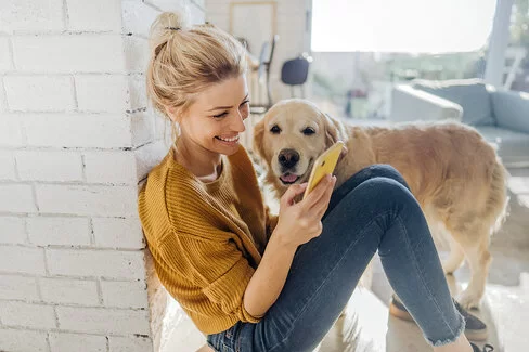 Junge Frau hockt in der Morgensonne an eine Mauer gelehnt auf dem Boden und liest etwas auf ihrem Smartphone, ein Hund steht an ihrer Seite und schaut ihr zu.