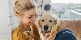 Junge Frau hockt in der Morgensonne an eine Mauer gelehnt auf dem Boden und liest etwas auf ihrem Smartphone, ein Hund steht an ihrer Seite und schaut ihr zu.