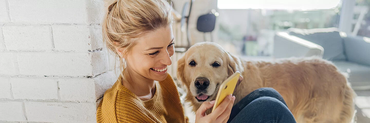 Junge Frau hockt in der Morgensonne an eine Mauer gelehnt auf dem Boden und liest etwas auf ihrem Smartphone, ein Hund steht an ihrer Seite und schaut ihr zu.