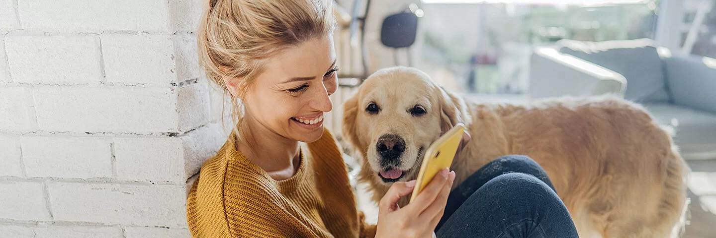 Junge Frau hockt in der Morgensonne an eine Mauer gelehnt auf dem Boden und liest etwas auf ihrem Smartphone, ein Hund steht an ihrer Seite und schaut ihr zu.