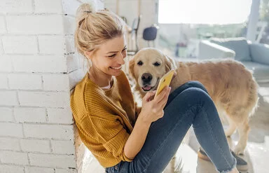Junge Frau hockt in der Morgensonne an eine Mauer gelehnt auf dem Boden und liest etwas auf ihrem Smartphone, ein Hund steht an ihrer Seite und schaut ihr zu.
