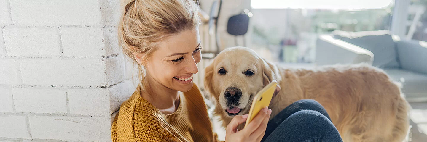 Junge Frau hockt in der Morgensonne an eine Mauer gelehnt auf dem Boden und liest etwas auf ihrem Smartphone, ein Hund steht an ihrer Seite und schaut ihr zu.