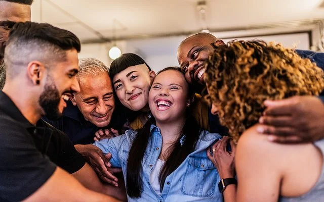 Eine Gruppe von sieben Menschen unterschiedlichen Alters, Geschlechts und Hautfarbe umarmt sich und lacht.