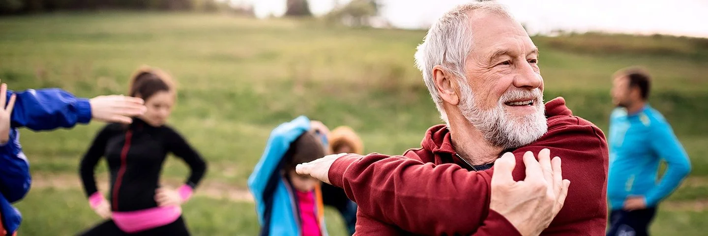 Älterer Mann in roter Jacke dehnt im Freien die Schultern, im Hintergrund wärmen sich weitere Teilnehmende einer Sportgruppe auf.