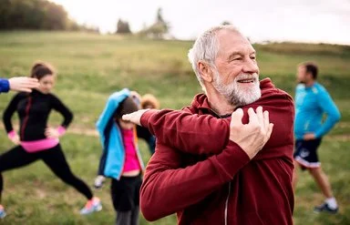 Älterer Mann in roter Jacke dehnt im Freien die Schultern, im Hintergrund wärmen sich weitere Teilnehmende einer Sportgruppe auf.