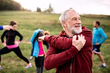 Älterer Mann in roter Jacke dehnt im Freien die Schultern, im Hintergrund wärmen sich weitere Teilnehmende einer Sportgruppe auf.