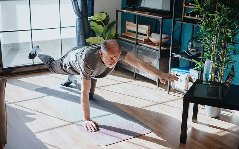 Ein älterer Mann führt im Wohnzimmer eine Gleichgewichtsübung aus. Er streckt dabei einen Arm und ein Bein von sich.