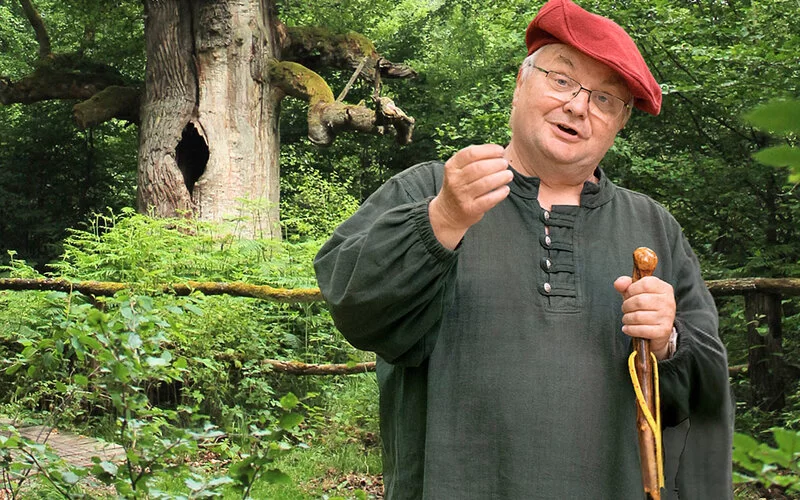 Älterer Mann in grünem Gewand und roter Mütze steht vor einem knorrigen Baum im Wald, hält einen Wanderstab und erläutert eine kleine Pflanze in seiner Hand.