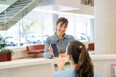 Eine lächelnde Frau in einem Jeanshemd zeigt ihr Smartphone bei der Anmeldung in einer Arztpraxis vor. Mit dem Smartphone zeigt sie eine Ersatzbescheinigung, welche anstatt ihrer Gesundheitskarte vorgezeigt werden kann