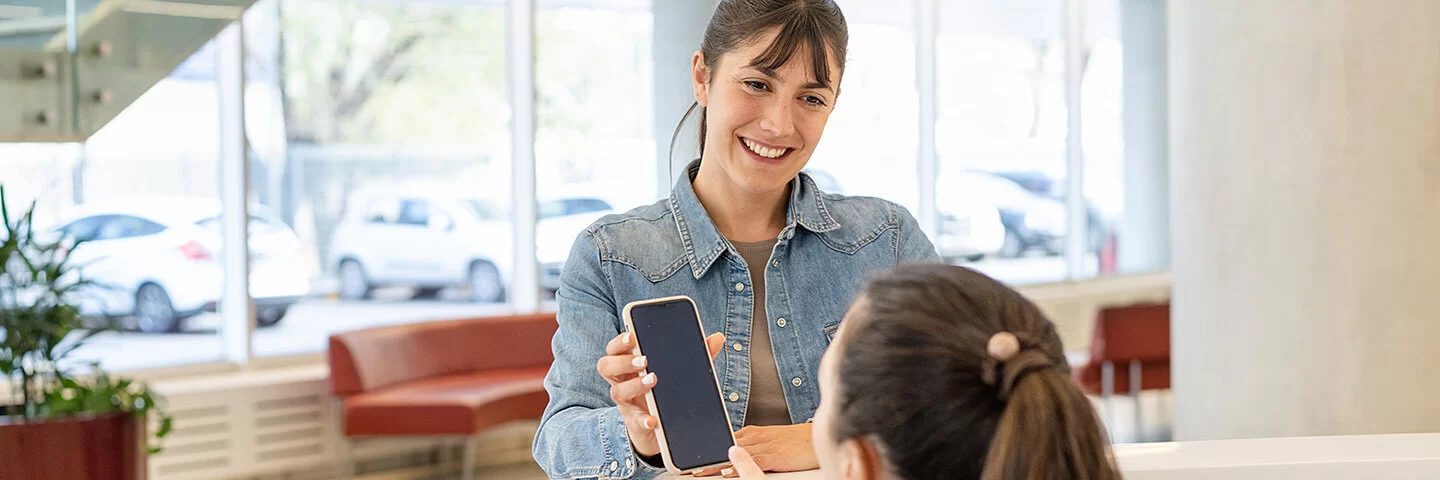 Eine lächelnde Frau in einem Jeanshemd zeigt ihr Smartphone bei der Anmeldung in einer Arztpraxis vor. Mit dem Smartphone zeigt sie eine Ersatzbescheinigung, welche anstatt ihrer Gesundheitskarte vorgezeigt werden kann