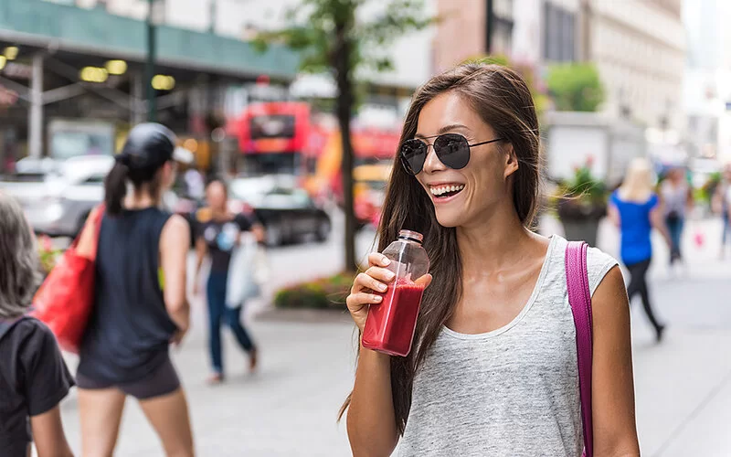 Eine junge Frau mit Sonnenbrille und kurzärmligem Top geht lächelnd durch eine Fußgängerzone und hält einen Smoothie in der Hand.