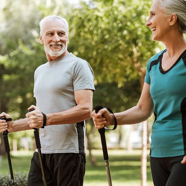 Mann und Frau, beide um die 50, in sportlicher Kleidung in einem Park. Sie halten Nordic-Walking-Stöcke in den Händen und lachen sich an.