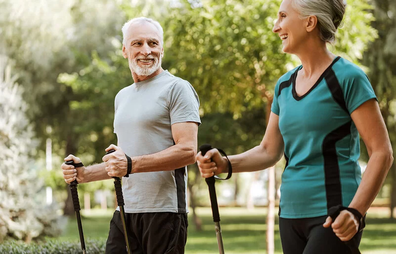 Mann und Frau, beide um die 50, in sportlicher Kleidung in einem Park. Sie halten Nordic-Walking-Stöcke in den Händen und lachen sich an.