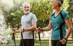 Mann und Frau, beide um die 50, in sportlicher Kleidung in einem Park. Sie halten Nordic-Walking-Stöcke in den Händen und lachen sich an.