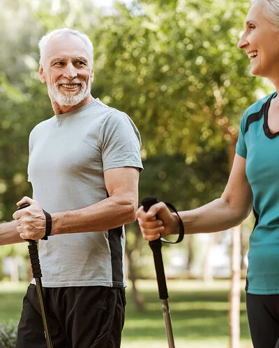 Mann und Frau, beide um die 50, in sportlicher Kleidung in einem Park. Sie halten Nordic-Walking-Stöcke in den Händen und lachen sich an.