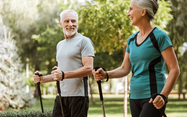 Mann und Frau, beide um die 50, in sportlicher Kleidung in einem Park. Sie halten Nordic-Walking-Stöcke in den Händen und lachen sich an.