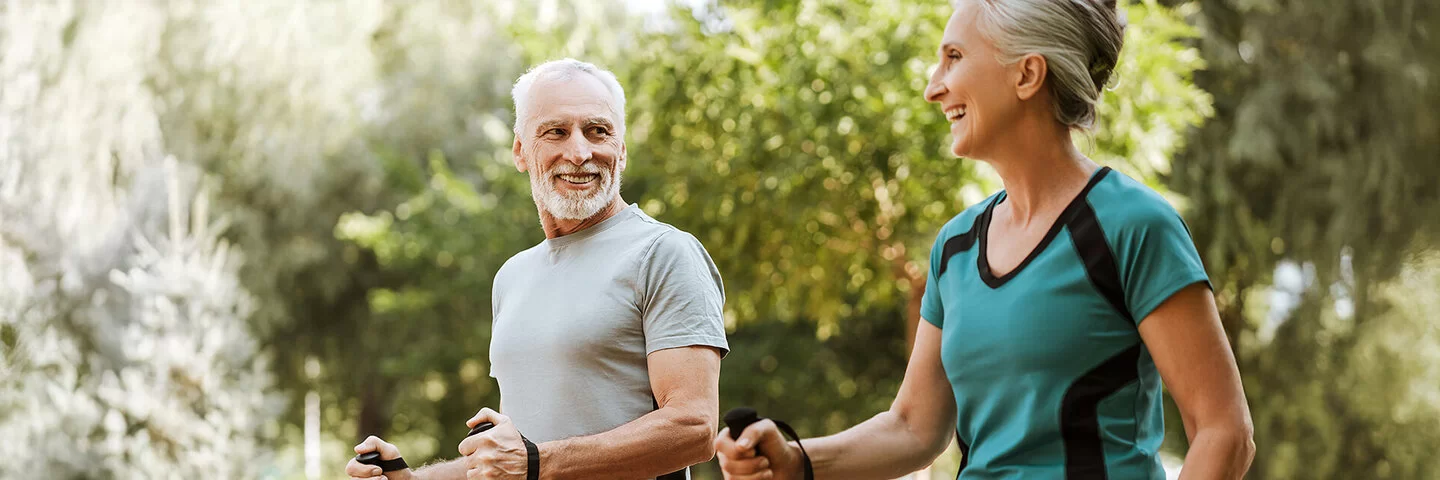 Mann und Frau, beide um die 50, in sportlicher Kleidung in einem Park. Sie halten Nordic-Walking-Stöcke in den Händen und lachen sich an.