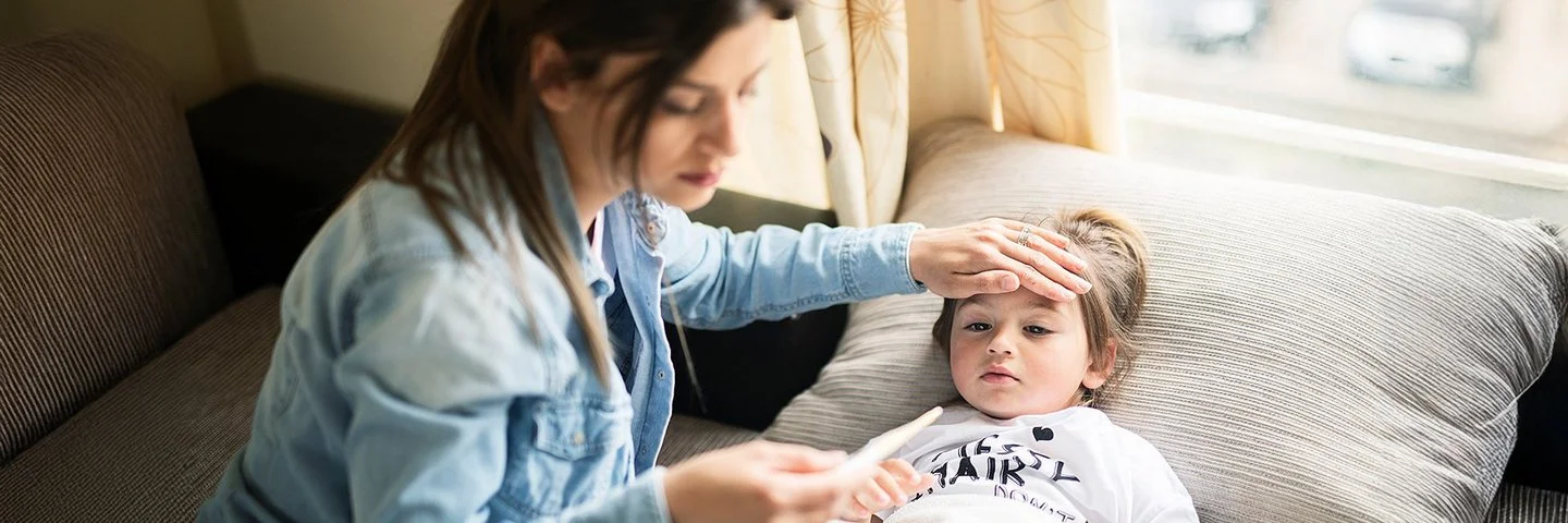 Eine Person mit langen, dunklen Haaren trägt eine Jeansjacke und sitzt auf einem Sofa. Die Hand liegt auf der Stirn eines Kindes, das auf dem Sofa unter einer weißen Decke liegt. In der anderen Hand hält die Person ein Fieberthermometer.