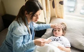 Eine Person mit langen, dunklen Haaren trägt eine Jeansjacke und sitzt auf einem Sofa. Die Hand liegt auf der Stirn eines Kindes, das auf dem Sofa unter einer weißen Decke liegt. In der anderen Hand hält die Person ein Fieberthermometer.