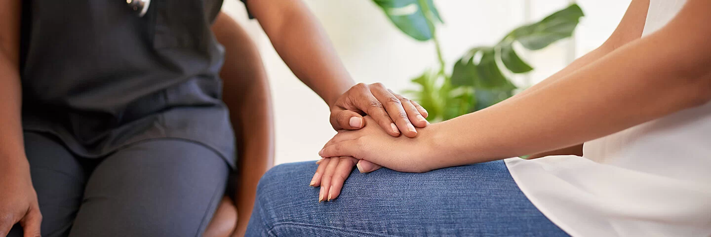 auf ihrem Knie. Eine Person in dunkler medizinischer Kleidung legt unterstützend eine Hand auf ihre. Die Szene vermittelt Trost und Unterstützung in einem medizinischen oder beratenden Umfeld. Im Hintergrund stehen grüne Pflanzen, die eine beruhigende Atmosphäre schaffen.