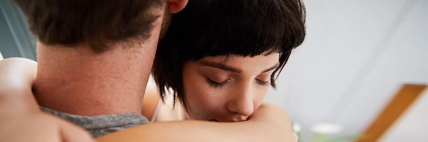 Eine junge Frau umarmt einen Mann. Ihre Augen sind geschlossen. Der Mann ist von hinten abgebildet, sein Gesicht ist nicht zu erkennen.