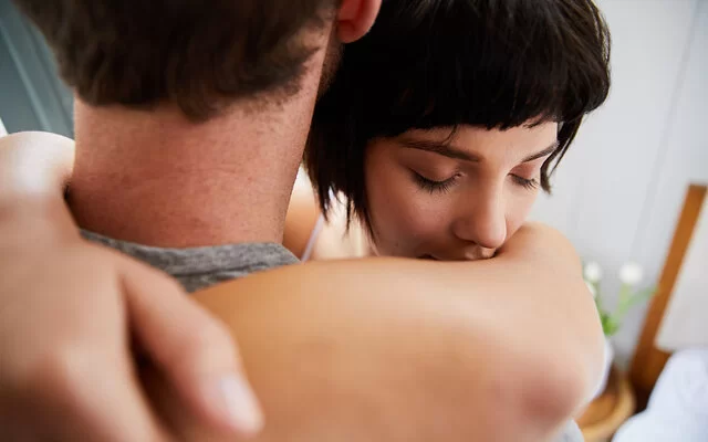 Eine junge Frau umarmt einen Mann. Ihre Augen sind geschlossen. Der Mann ist von hinten abgebildet, sein Gesicht ist nicht zu erkennen.