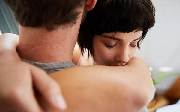 Eine junge Frau umarmt einen Mann. Ihre Augen sind geschlossen. Der Mann ist von hinten abgebildet, sein Gesicht ist nicht zu erkennen.