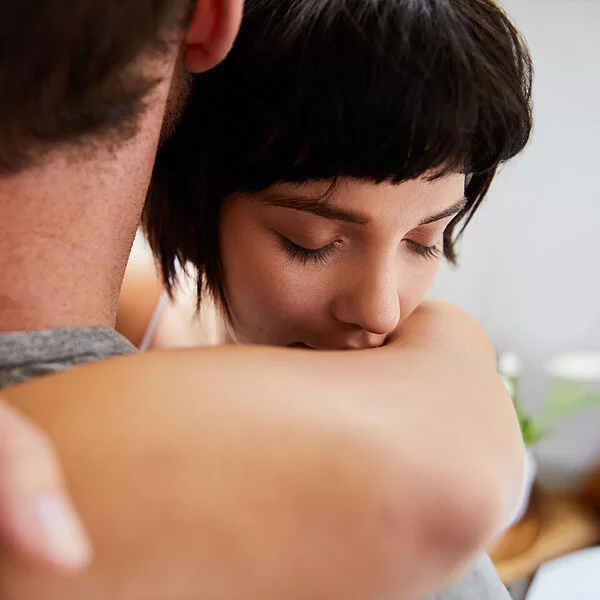 Eine junge Frau umarmt einen Mann. Ihre Augen sind geschlossen. Der Mann ist von hinten abgebildet, sein Gesicht ist nicht zu erkennen.