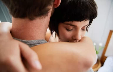 Eine junge Frau umarmt einen Mann. Ihre Augen sind geschlossen. Der Mann ist von hinten abgebildet, sein Gesicht ist nicht zu erkennen.