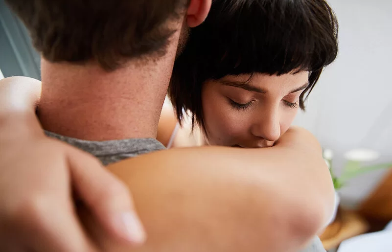 Eine junge Frau umarmt einen Mann. Ihre Augen sind geschlossen. Der Mann ist von hinten abgebildet, sein Gesicht ist nicht zu erkennen.
