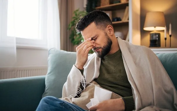 Ein Mann sitzt auf dem Sofa und fasst sich mit der rechten Hand an die Nasenwurzel, in der linken hält er ein Taschentuch.