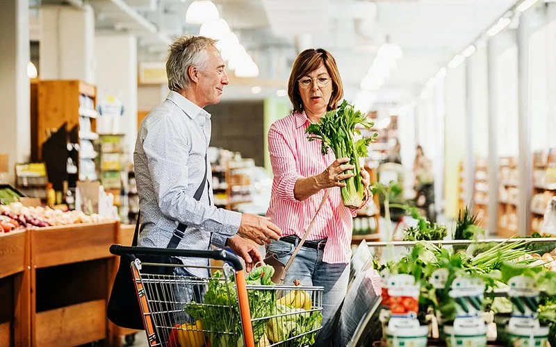 Ein Paar kauft Gemüse im Supermarkt. Die Frau hält einen Bund Sellerie, ihr Mann steht neben dem Einkaufswagen.