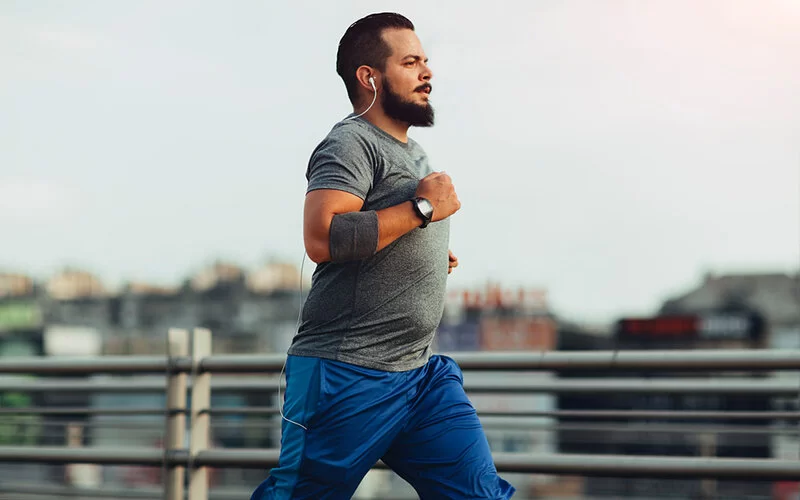 Ein übergewichtiger junger Mann joggt in einer Großstadt über eine Brücke.