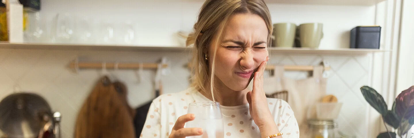 Eine Frau steht mit einem Glas Wasser in der Hand in ihrer Küche. Mit schmerzverzerrtem Gesichtsausdruck hält sie sich die Hand an die Wange.