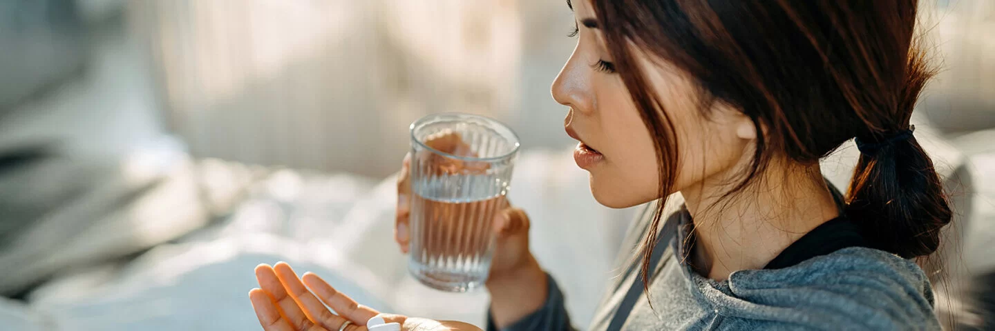 Eine junge Frau sitzt aufrecht im Bett, in der einen Hand hält sie ein Glas Wasser und in der anderen Handfläche liegen drei weiße Tabletten, die sie einnehmen möchte.