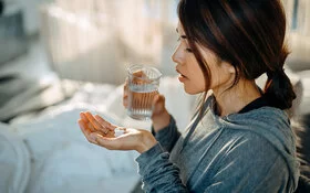 Eine junge Frau sitzt aufrecht im Bett, in der einen Hand hält sie ein Glas Wasser und in der anderen Handfläche liegen drei weiße Tabletten, die sie einnehmen möchte.