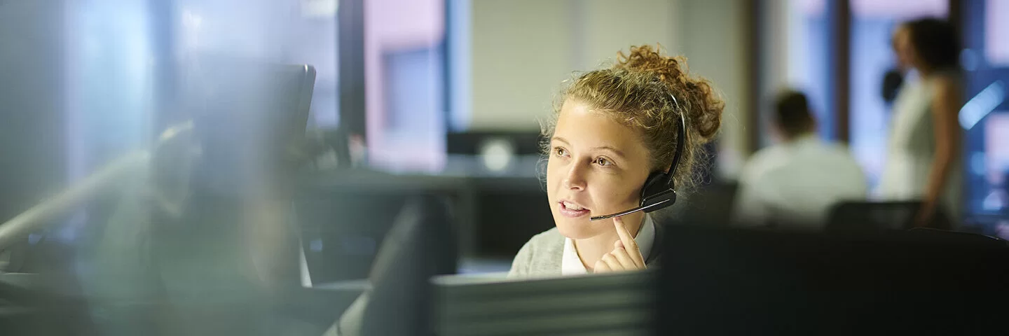 Eine Frau sitzt mit einem Headset vor einem Computerbildschirm und telefoniert. 