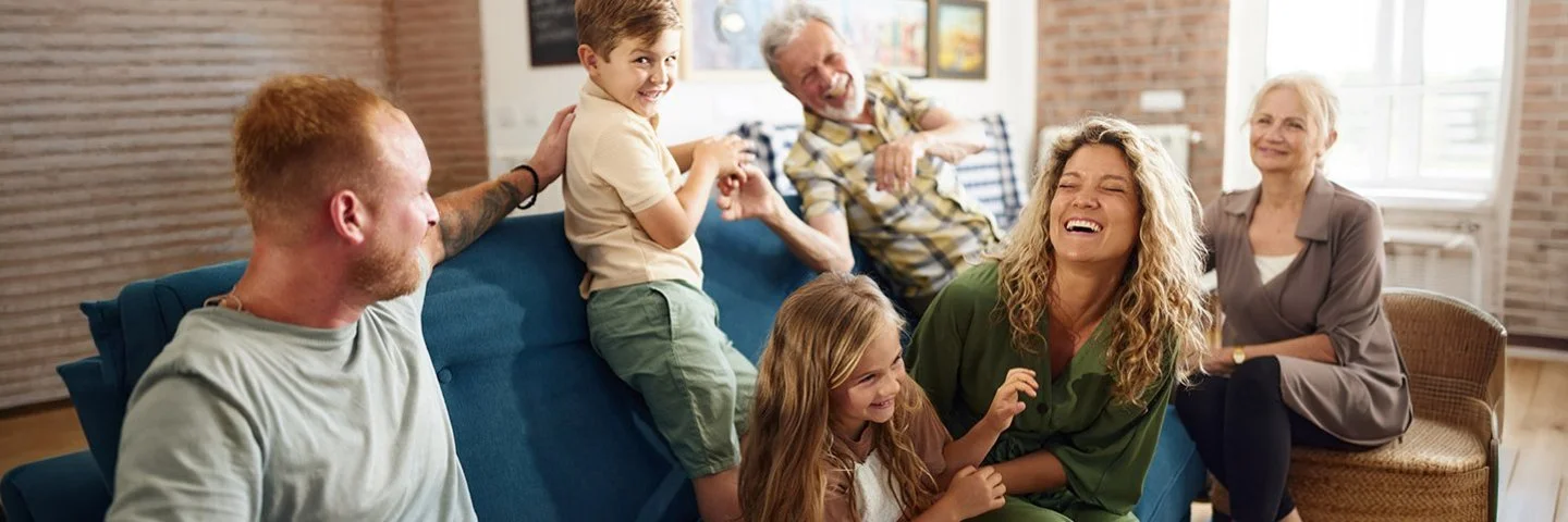 Sechs Personen unterschiedlichen Alters sitzen in einem Wohnzimmer, lachen und unterhalten sich entspannt auf einem Sofa und einem Sessel