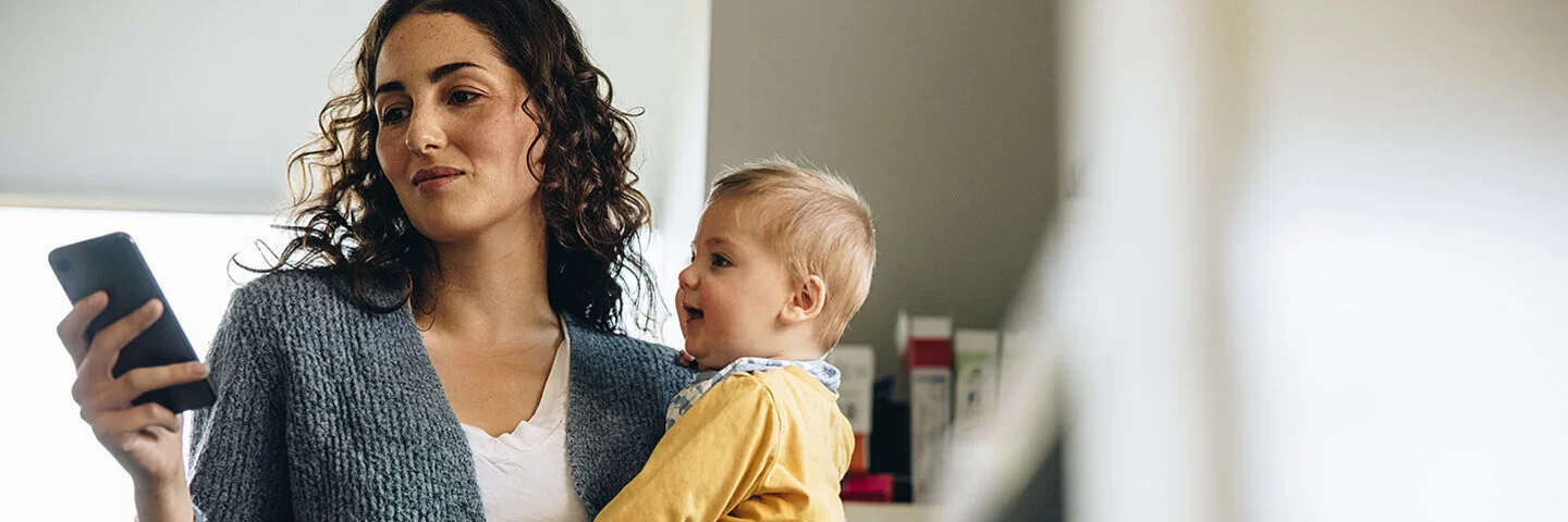 Eine junge Mutter steht in der Küche und hält ihr Baby auf dem Arm. In der rechten Hand hält sie ein Smartphone und liest sich darauf etwas durch.