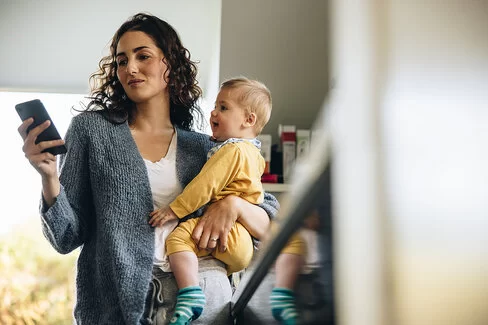 Eine junge Mutter steht in der Küche und hält ihr Baby auf dem Arm. In der rechten Hand hält sie ein Smartphone und liest sich darauf etwas durch.