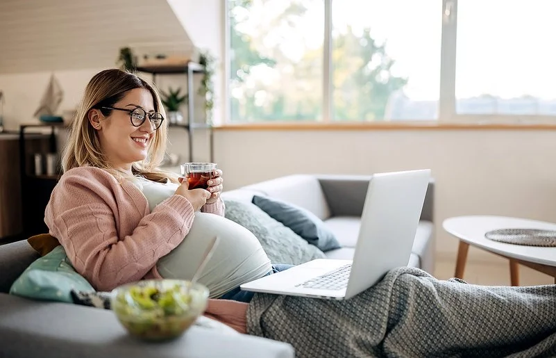 Eine schwangere Frau sitzt mit einem Glas Tee auf dem Sofa und lächelt.
