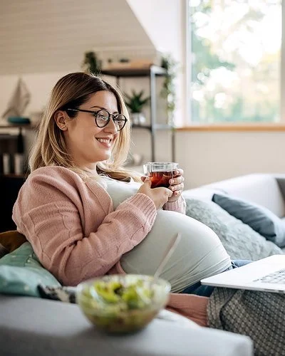 Eine schwangere Frau sitzt mit einem Glas Tee auf dem Sofa und lächelt.