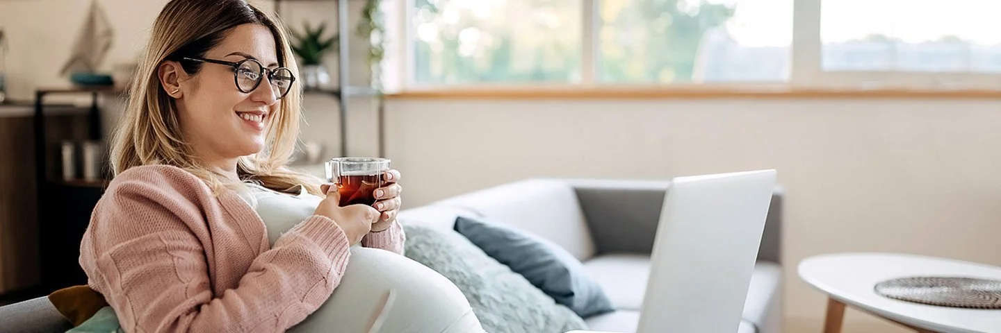 Eine schwangere Frau sitzt mit einem Glas Tee auf dem Sofa und lächelt.