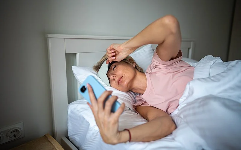 Eine Frau nimmt eine Schlafmaske ab und sieht auf ihr Handy.