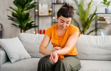 Eine junge Frau in einem Wohnzimmer. Sie sitzt auf einem Sofa und betrachtet die Innenseite ihres linken Unterarms. Dort ist ein schwach roter Strich auf der Haut zu sehen, der sich vom Handgelenk bis zum Ellenbogen zieht. Ihr Blick ist leicht besorgt.