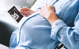 Eine schwangere Frau sitzt auf dem Sofa, hält ihren Bauch und betrachtet ein Ultraschall-Bild.