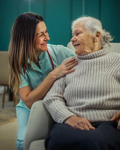 Eine alte Frau sitzt in einem Sessel. Neben ihr kniet eine Pflegekraft und legt ihre Hände auf die Schultern der Frau. Sie sehen sich in die Augen und lächeln.