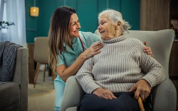 Eine alte Frau sitzt in einem Sessel. Neben ihr kniet eine Pflegekraft und legt ihre Hände auf die Schultern der Frau. Sie sehen sich in die Augen und lächeln.
