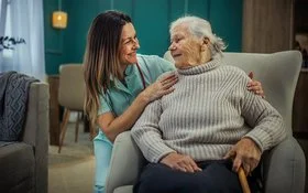 Eine alte Frau sitzt in einem Sessel. Neben ihr kniet eine Pflegekraft und legt ihre Hände auf die Schultern der Frau. Sie sehen sich in die Augen und lächeln.